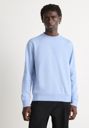 Ung man i ljusblå crewneck-tröja med "BOSS"-logotyp och svarta byxor, som står mot en enfärgad vit bakgrund.