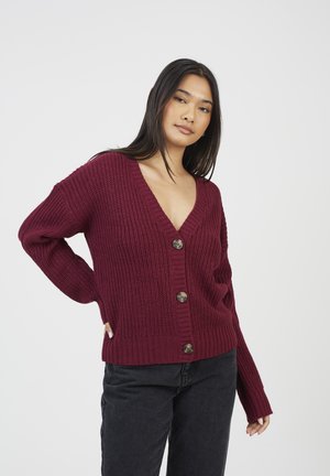 Vrouw in een bordeauxrode V-hals gebreide cardigan met grote knopen en zwarte broek, poseert met één hand op haar heup tegen een witte achtergrond.