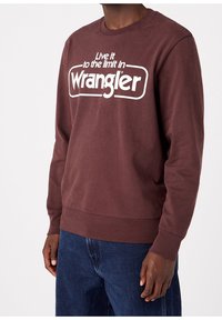 Uomo che indossa una felpa bordeaux con il testo bianco "Vivi al limite con Wrangler" e jeans blu, in piedi contro uno sfondo semplice.