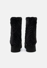 Pár černých, prošívaných kotníkových bot z umělé kožešiny s vysokými platformovými podpatky zobrazených zezadu na bílém pozadí.