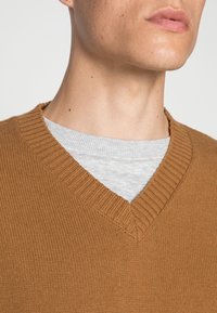 Brun v-ringad tröja med ribbad krage, överlagd på en grå skjorta. Tröjan har ett strukturerat stickat mönster. Närbild av överkroppen.