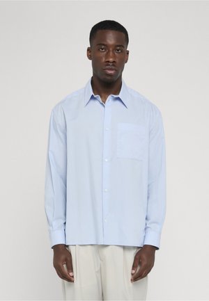 Mand iført lyseblå langærmet skjorte med knapper og lyse beige bukser, stående mod en ensfarvet lys baggrund.
