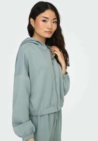 Lys grønne zip-up hettegenser laget av myk stoff, med snøring i livet og elastiske mansjetter, med en avslappet passform og hette.