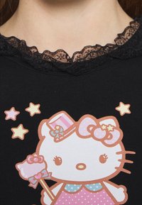 Dítě oblečené v černém tričku s krajkovým výstřihem, na kterém je bílá kreslená kočka držící hůlku, obklopená žlutými a růžovými hvězdami.