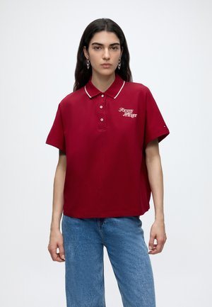 Kvinde med langt mørkt hår iført en oversized rød polo med "Tommy Hilfiger" logo og lyseblå jeans mod en ensfarvet baggrund.