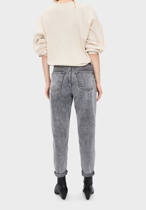 Personne debout de dos, portant un haut à manches longues beige clair, un jean gris délavé roulé aux chevilles, et des bottines noires à talons.