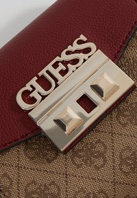 Bordeaux syntetisk læder håndtaske med guldfarvet hardware, der har en struktureret brun stofbund og et fremtrædende brandlogo på klaffen.