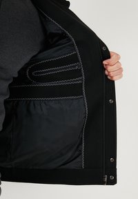 Juodas atvertas švarkas, kuriame matyti lygi tamsi pamušalas ir kelios vidinės kišenės su kontrastingais baltais siūlais.