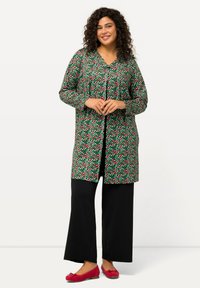 Cappotto lungo con bottoni su sfondo verde, caratterizzato da motivi a pois rossi e rosa. Abbinato a pantaloni a gamba larga neri e ballerine rosse.