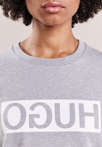 Sudadera gris con un gran gráfico blanco que muestra la palabra "HUGO" en letras gruesas y modernas. Material de suave mezcla de algodón, diseño de cuello redondo.