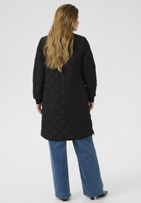 Manteau long noir matelassé avec fentes sur les côtés et manches longues, porté sur un jean large bleu, dotée d'une texture lisse et d'un design simple.