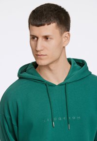 Grön hoodie i mjukt material med känguruficka, justerbar dragsko och broderad text "LINDENBERGH" på framsidan.