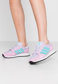 Des chaussures de sport violettes et roses en maille et en daim, avec trois bandes teal, une semelle intermédiaire blanche et une semelle extérieure en caoutchouc noir.