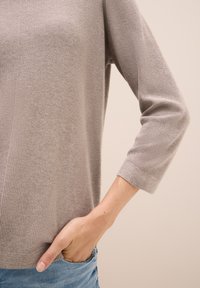 Hellbrauner, gestrickter Pullover mit strukturierter Oberfläche, Dreiviertelärmeln und lockerer Passform. Hand in einer blauen Jeansentasche.