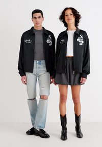 Črne zip-up jakne z vezenimi grafičnimi motivi, oblečene čez sivo srajco in belo majico. En model v obrabljenih kavbojkah, drugi v nabranem krilu.