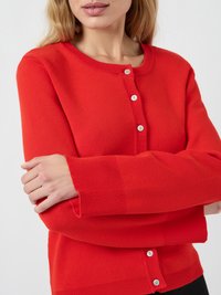 Frau mit heller Haut und blonden Haaren, die einen leuchtend roten Knopf-Cardigan trägt und die Arme über der Brust verschränkt hat.