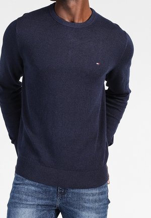 Hombre vestido con un suéter azul marino de cuello redondo con un pequeño logo en el pecho y jeans azules, de pie con las manos detrás de la espalda.