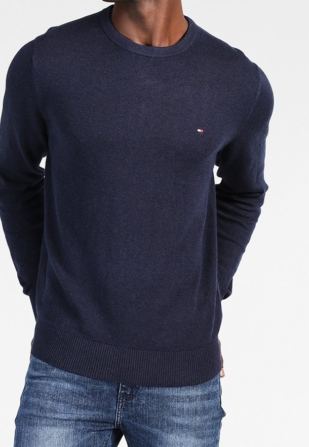 Homme portant un pull ras-du-cou bleu marine avec un petit logo sur la poitrine et un jean bleu, debout les mains derrière le dos.
