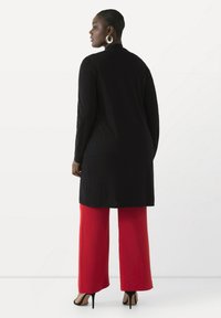 Langer schwarzer Cardigan mit geradem Schnitt, kombiniert mit roten weiten Hosen, die eine glatte Textur und ein einfaches Design aufweisen.