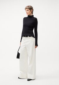 Pull à col roulé noir côtelé, associé à un large pantalon blanc. Un sac à main noir et des chaussures noires à talons pointus complètent la tenue. Design épuré et moderne.