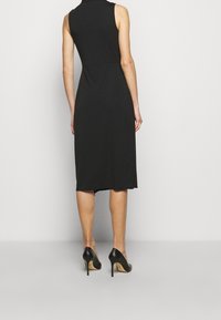 Svart ärmlös klänning med figurnära midja, knälång fåll och mjuk textur, kombinerad med svarta högklackade skor.