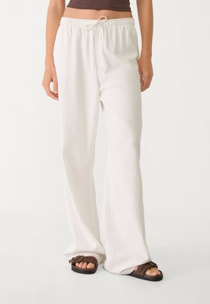 Mujer que lleva puestos unos pantalones blancos holgados con cordón y sandalias marrones de punta abierta, de pie frente a un fondo liso.