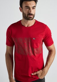 Rode katoenen t-shirt met korte mouwen, voorzien van een pocket en een gestructureerd grafisch ontwerp in grijs op de voorkant. Ronde halslijn.