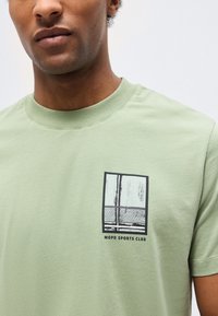 Mees, kes kannab helerohelise T-särgi peal must-valge spordivõrgu graafika ja rinnal tekstiga "MOPD SPORTS CLUB".