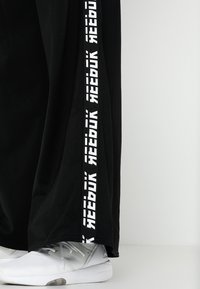 Pantalones deportivos negros con una franja blanca vertical de marca Reebok a lo largo del costado, combinados con zapatillas deportivas blancas.