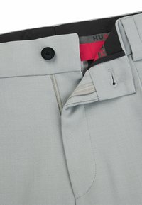 Light gray trousers with a textured fabric, button closure, and zipper fly. Features a contrasting red inner waistband and precise stitching.