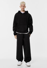 Fekete kötött kapucnis pulóver elülső zsebbel, laza fekete melegítőnadrággal, amelyet zsinóros derék jellemez. A fehér sportcipő kiegészíti a szettet.