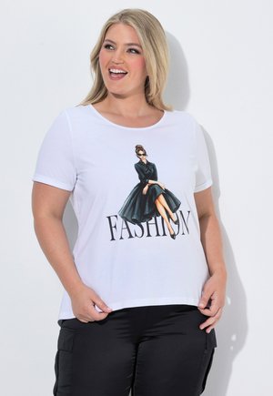 Uśmiechnięta kobieta o blond włosach nosi białą koszulkę z ilustracją siedzącej kobiety i słowem "FASHION" na czarnych spodniach.