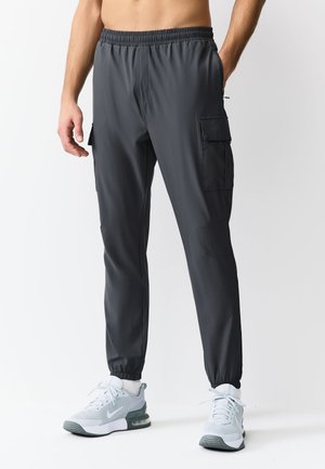 Man wearing dark gray cargo jogger pants and white athletic sneakers, standing against a plain white background.