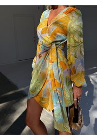 Femme portant une robe portefeuille colorée avec des motifs de feuilles jaunes, vertes et bleues, tenant un sac à main beige tissé avec une anse noire.