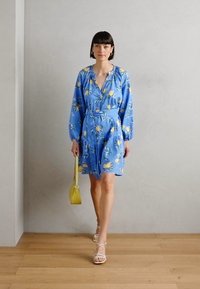 Rochie florală albastră cu mâneci lungi, talie strânsă și model de lămâie. Asortată cu sandale albe cu bretele și o geantă mică galbenă.