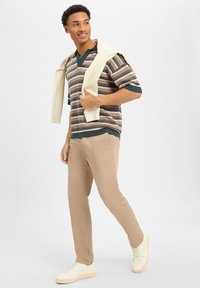 Bruin en teal gestreepte gebreide polo trui, beige chinos, een crèmekleurige trui vasthoudend, en beige sneakers. Casual outfit met gelaagde texturen.