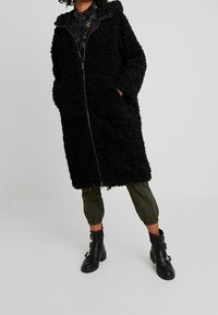 Abrigo negro, esponjoso y de largo con cierre de cremallera, bolsillos laterales y capucha. Llevado sobre pantalones verde olivo y combinado con botas negras de hebilla.