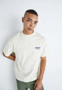 Cream short-sleeve t-shirt with "ORIGINALS" printed in blue on the left chest. Soft cotton fabric with relaxed fit, paired with green pants.