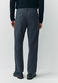 Osoba w ciemnoszarych spodniotablicowych w kratę z zapiętymi tylnymi kieszeniami, czarnych butach i ciemnej bluzie z długim rękawem, stoi na jednolitym tle.