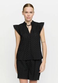 Svart blus i ögla med nyckelhålskrage, volangärmar och scalloped nederkant, i kombination med matchande shorts i samma material.