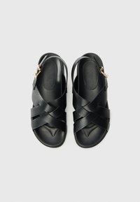 Black sandals with a cross strap design, made of smooth material, featuring an adjustable ankle strap with gold hardware accents.