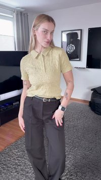 Άτομο με μακριά ξανθά μαλλιά, ντυμένο με μπεζ υφασμένη κοντομάνικη μπλούζα πόλο και μαύρο παντελόνι, στέκεται σε μοντέρνο σαλόνι με ορατά τατουάζ.