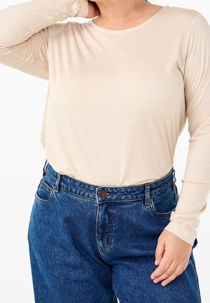 Personne portant une chemise beige à manches longues rentrée dans un jean en denim bleu taille haute, avec une main reposant sur la hanche.