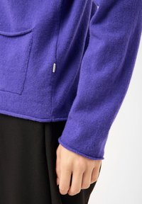 Nærbillede af en persons hånd nær sidesømmen på en lilla strikket trøje med en lille sidelomme og sorte bukser.