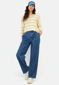 Helles gelbes und weiß gestreiftes Langarmoberteil, blaue hochgeschnittene, weit geschnittene Jeans und weiße Sneaker. Eine Denim-Cap rundet den Look ab.