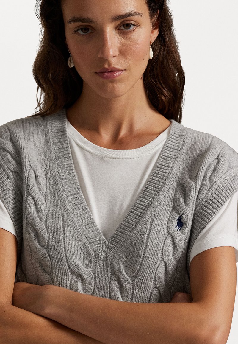 Grå kabelstrikket ærmeløs sweater over en hvid skjorte, med en dyb V-udskæring og et lille marineblåt logo på venstre side.