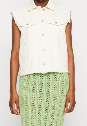 Veste en denim blanche sans manches avec fermeture à boutons et bords effilochés portée sur une jupe tricotée vert clair.