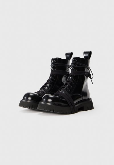 Bottes de combat en cuir noir avec des lacets hauts, une semelle en caoutchouc épaisse et des sangles en tissu enveloppées autour de la cheville. Bout rond et détail de tirette.