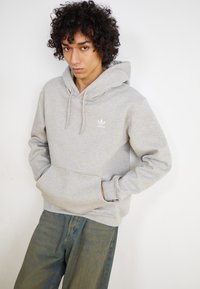 Pelēka kapuce krekli ar kangaroo kabatu, faktūras audums un balta adidas logo uz krūtīm. Apvienots ar brīvām zilo džinsu biksēm.