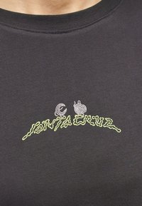 Κοντινό πλάνο σκούρου t-shirt με κίτρινο-πράσινο κυματιστό κείμενο "Santa Cruz" και δύο μικρά λευκά σπιράλ σχέδια πάνω από τα γράμματα.